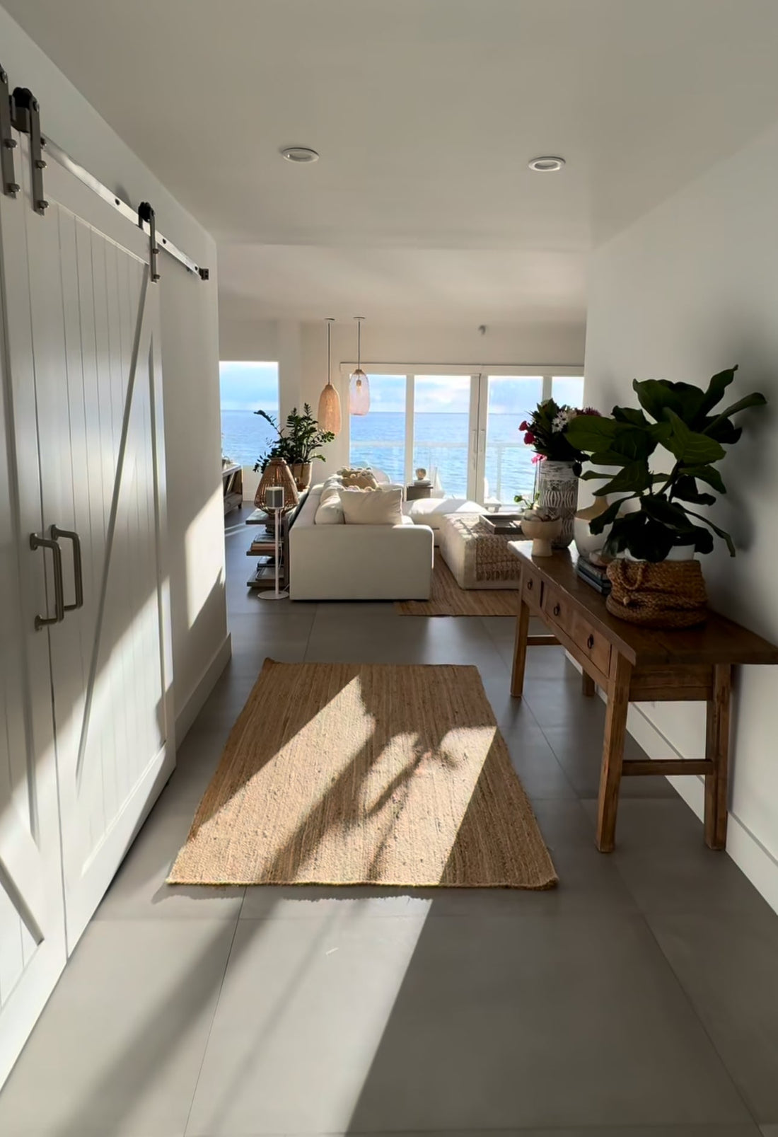 Bright coastal entryway leading into a living room with a white sofa, natural wood console table, woven rugs, indoor plants, and large windows overlooking the ocean